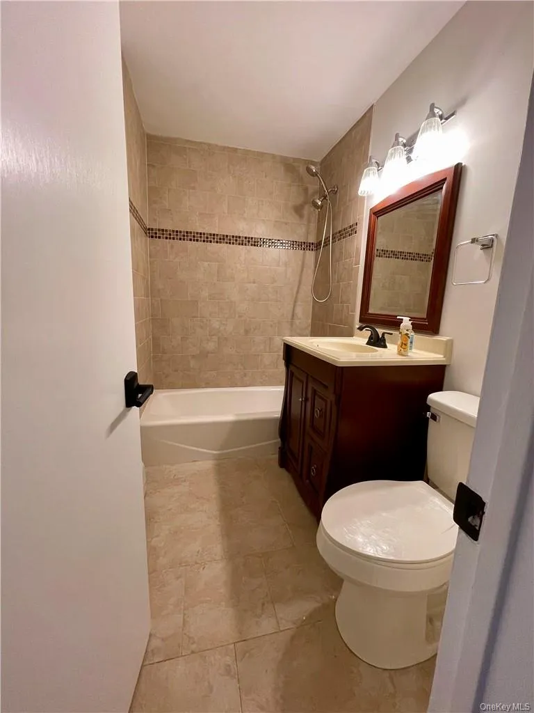 Bathroom featuring a white bathtub, tiled shower surround with decorative accent, single vanity with wood-finish cabinetry, countertop, and a wall-mounted mirror with overhead lighting Bathroom featuring a white bathtub, tiled shower surround with decorative accent, single vanity with wood-finish cabinetry, countertop, and a wall-mounted mirror with overhead lighting