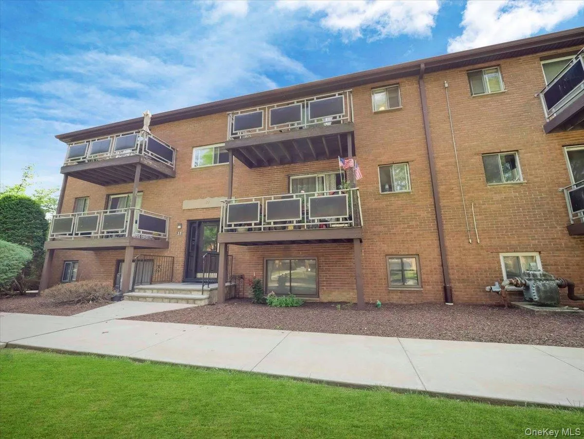 Multi-level brick exterior building featuring individual balconies with metal railings, a central entry door, mature shrubbery, and a well-maintained lawn Multi-level brick exterior building featuring individual balconies with metal railings, a central entry door, mature shrubbery, and a well-maintained lawn