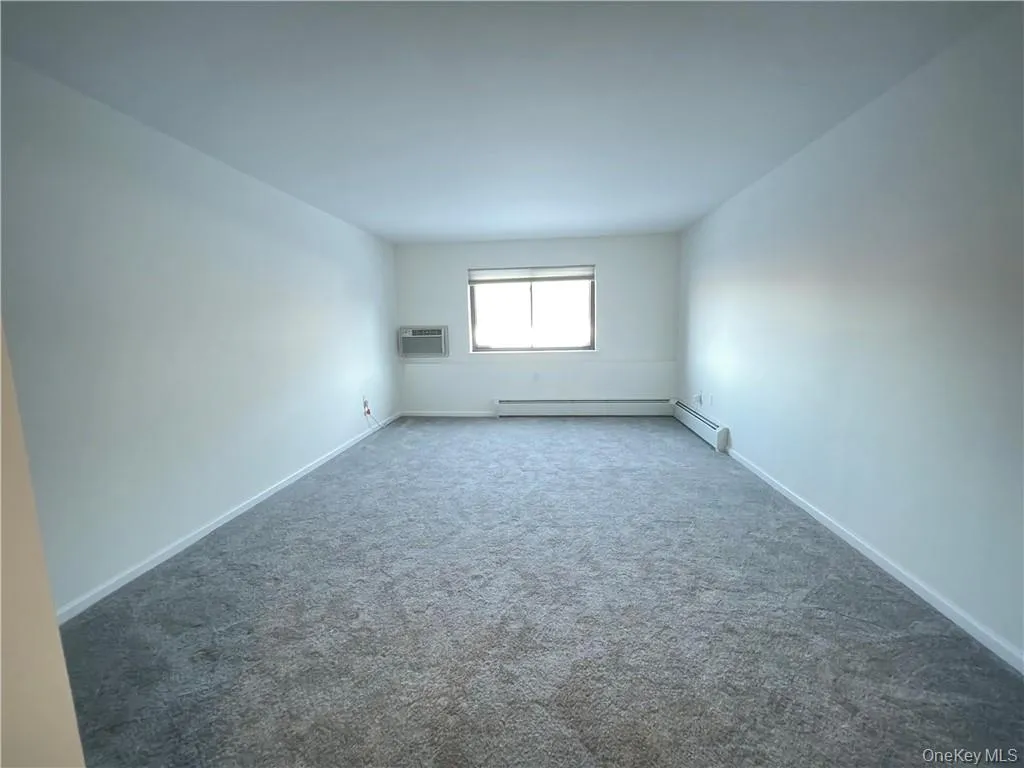Spacious room featuring gray carpeting, white walls, a window with natural light, a window-mounted air conditioning unit, and baseboard heating Spacious room featuring gray carpeting, white walls, a window with natural light, a window-mounted air conditioning unit, and baseboard heating