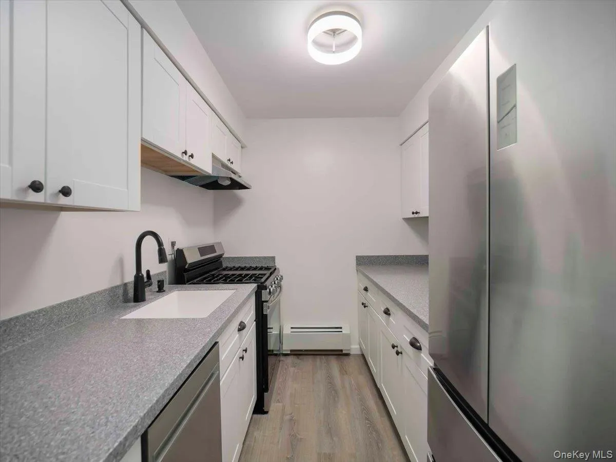 Galley kitchen featuring white shaker cabinetry, speckled countertops, stainless steel appliances, and wood-finish flooring Galley kitchen featuring white shaker cabinetry, speckled countertops, stainless steel appliances, and wood-finish flooring