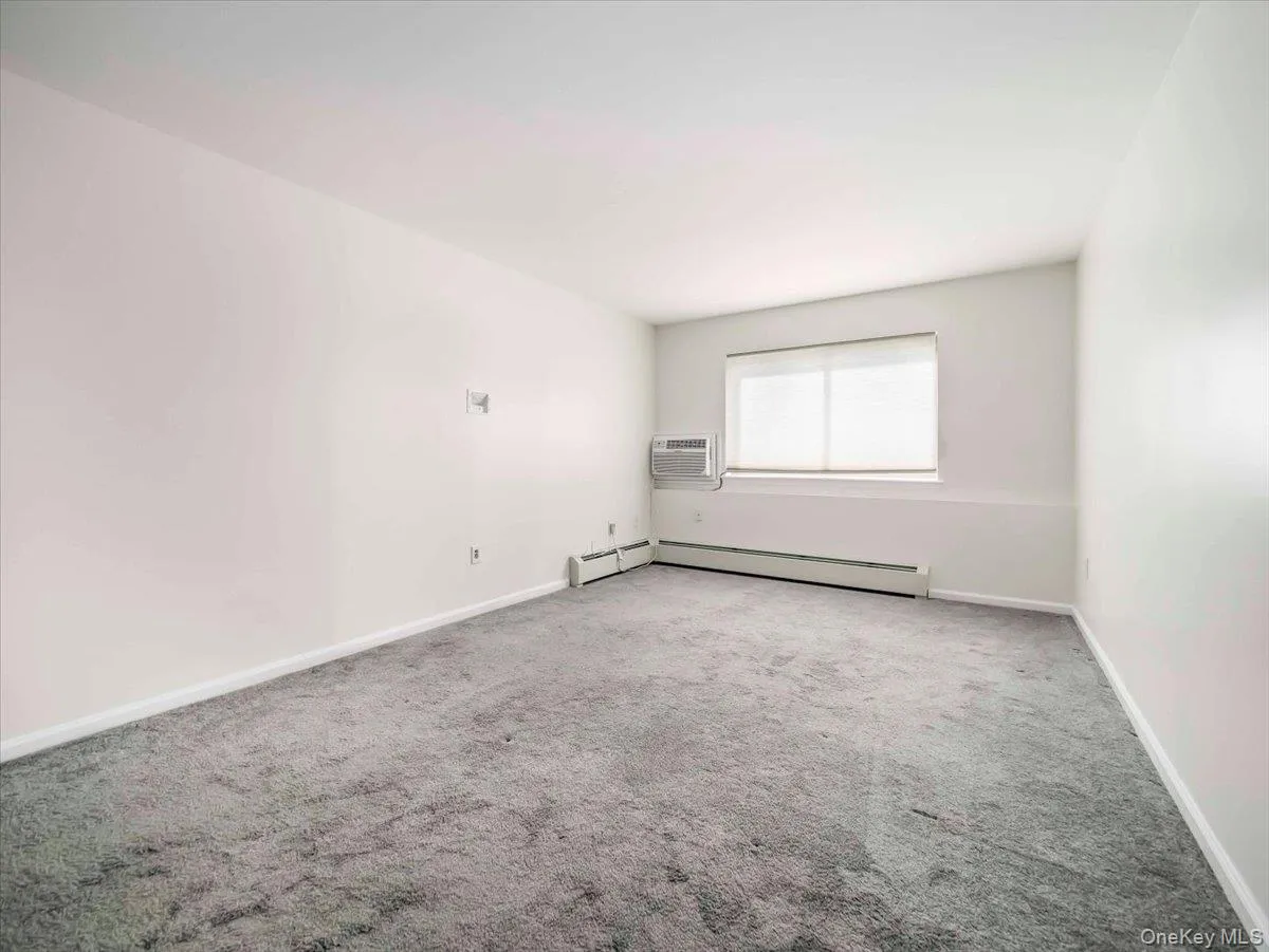 Neutral wall paint, gray carpeting, a window with horizontal blinds, and a wall-mounted air conditioning unit Neutral wall paint, gray carpeting, a window with horizontal blinds, and a wall-mounted air conditioning unit