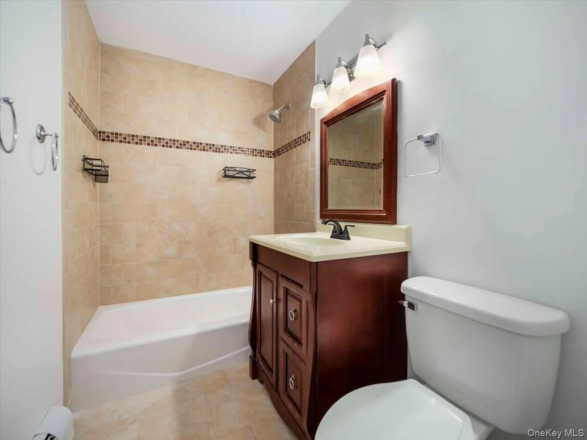 Bathroom featuring a tiled shower surround with decorative accent band, a white bathtub, and a wood-finish vanity with an integrated sink Bathroom featuring a tiled shower surround with decorative accent band, a white bathtub, and a wood-finish vanity with an integrated sink