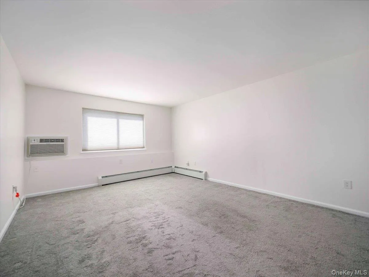 Carpeted interior space with a window, window-mounted air conditioning unit, and baseboard heating Carpeted interior space with a window, window-mounted air conditioning unit, and baseboard heating