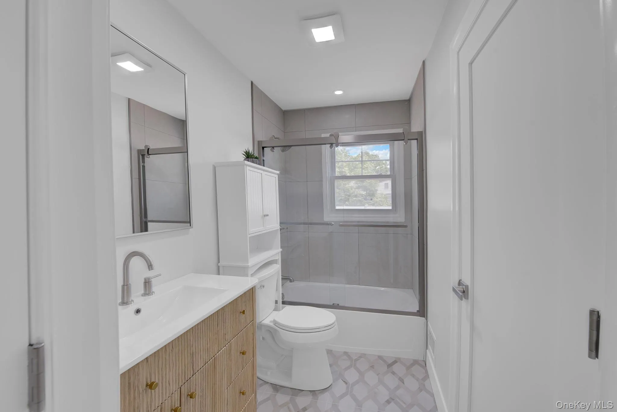 Bathroom featuring a wood-finish vanity with an integrated sink, a frameless mirror, a built-in bathtub with a sliding glass enclosure, recessed lighting, and patterned floor tile Bathroom featuring a wood-finish vanity with an integrated sink, a frameless mirror, a built-in bathtub with a sliding glass enclosure, recessed lighting, and patterned floor tile