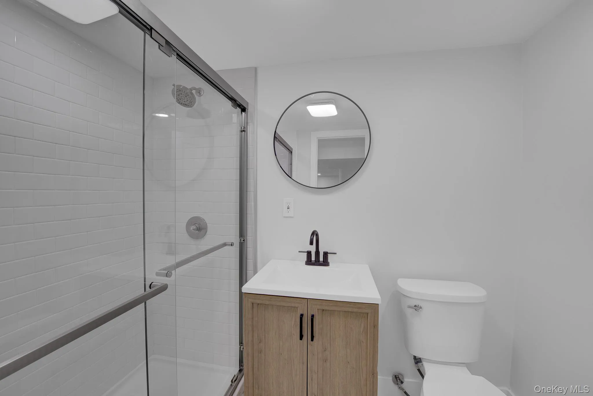 Bathroom featuring a glass-enclosed shower with subway tile surround, a circular vanity mirror, a wood-finish vanity with a white countertop, and a matte black faucet Bathroom featuring a glass-enclosed shower with subway tile surround, a circular vanity mirror, a wood-finish vanity with a white countertop, and a matte black faucet