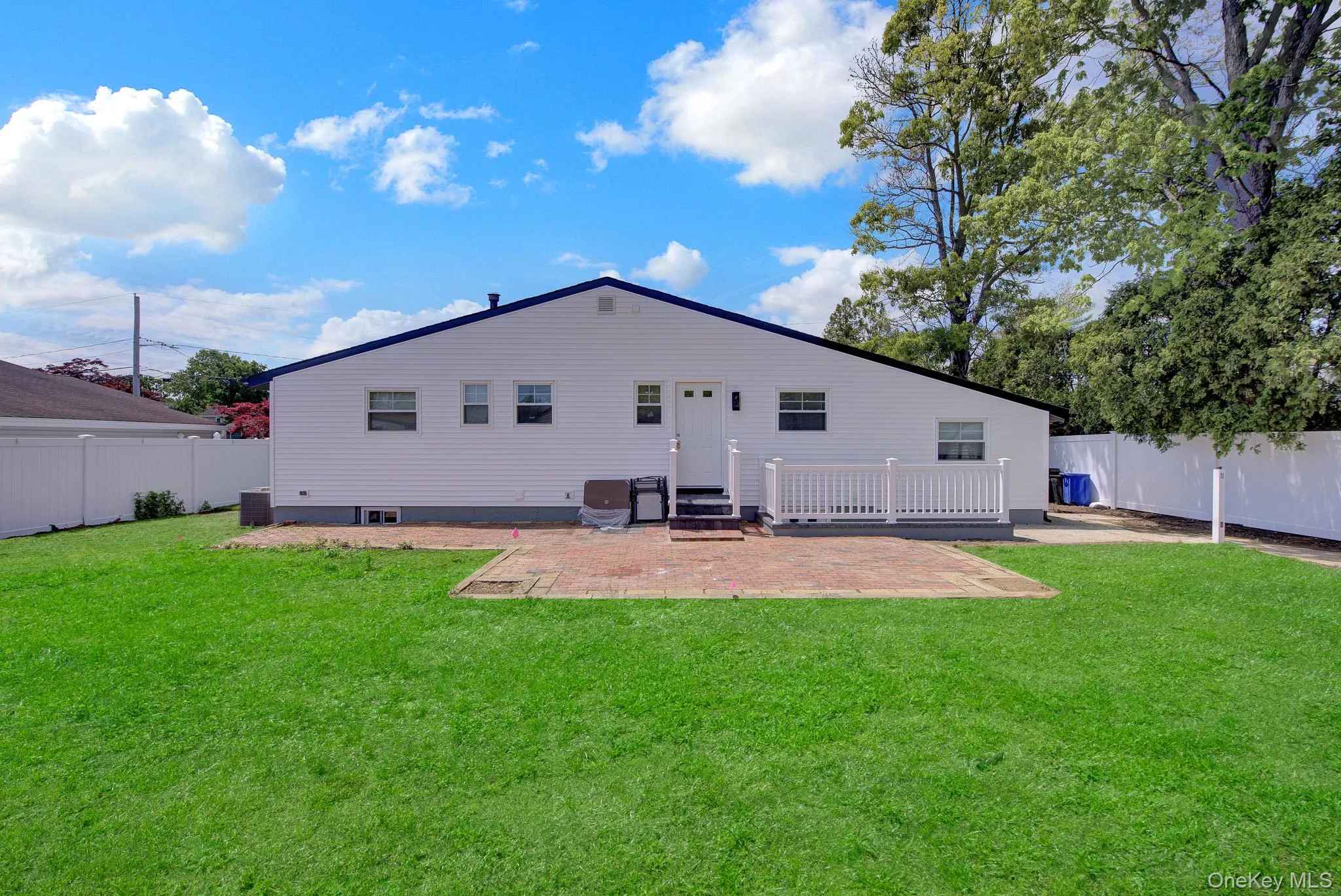 Spacious backyard featuring a large green lawn, a brick paver patio, white vinyl fencing, and a white deck with railings Spacious backyard featuring a large green lawn, a brick paver patio, white vinyl fencing, and a white deck with railings