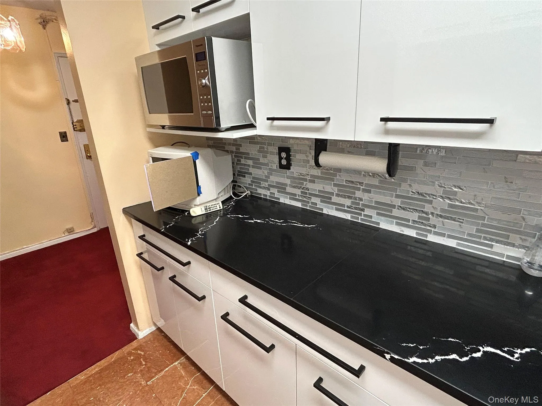 Modern kitchen area featuring heavy black quartz countertops with white veining, white cabinetry with black bar pulls, and a mosaic tile backsplash Modern kitchen area featuring heavy black quartz countertops with white veining, white cabinetry with black bar pulls, and a mosaic tile backsplash