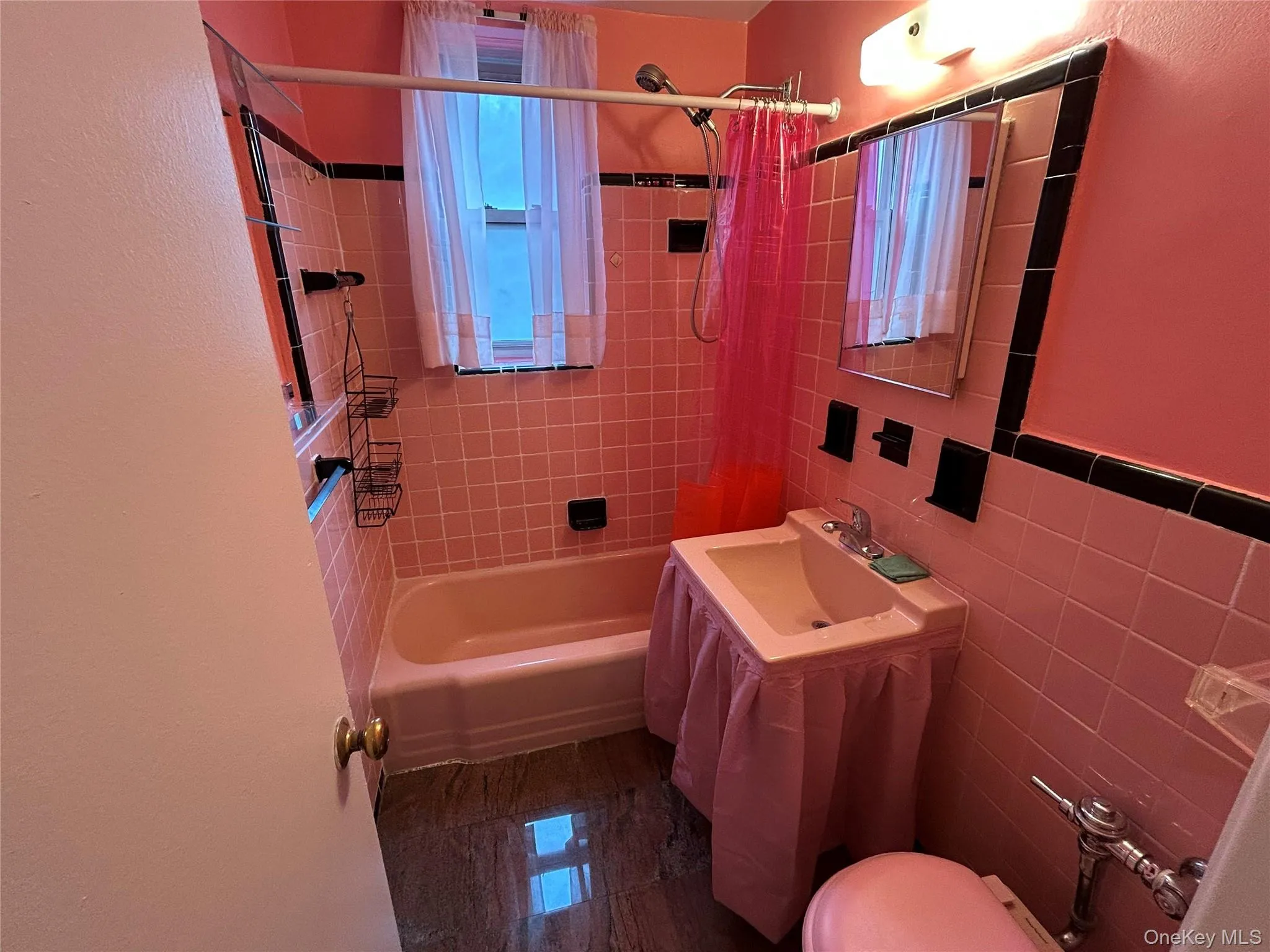 Bathroom featuring pink tiled walls with black accent trim, a built-in pink bathtub, a window with natural light, a wall-mounted sink, and a built-in mirror Bathroom featuring pink tiled walls with black accent trim, a built-in pink bathtub, a window with natural light, a wall-mounted sink, and a built-in mirror