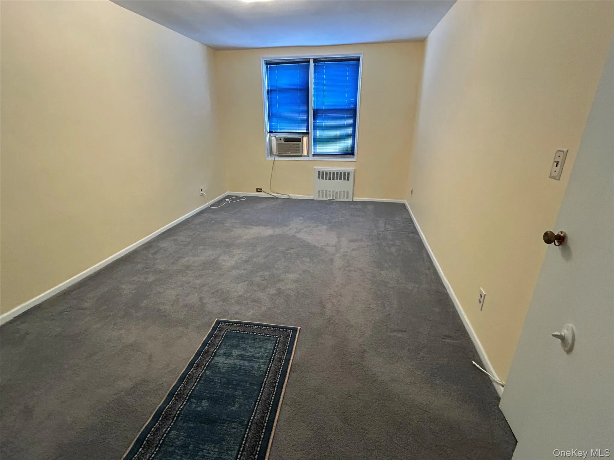 Carpeted bedroom room with a window-mounted air conditioning unit, baseboard heating, and light-toned walls Carpeted bedroom room with a window-mounted air conditioning unit, baseboard heating, and light-toned walls