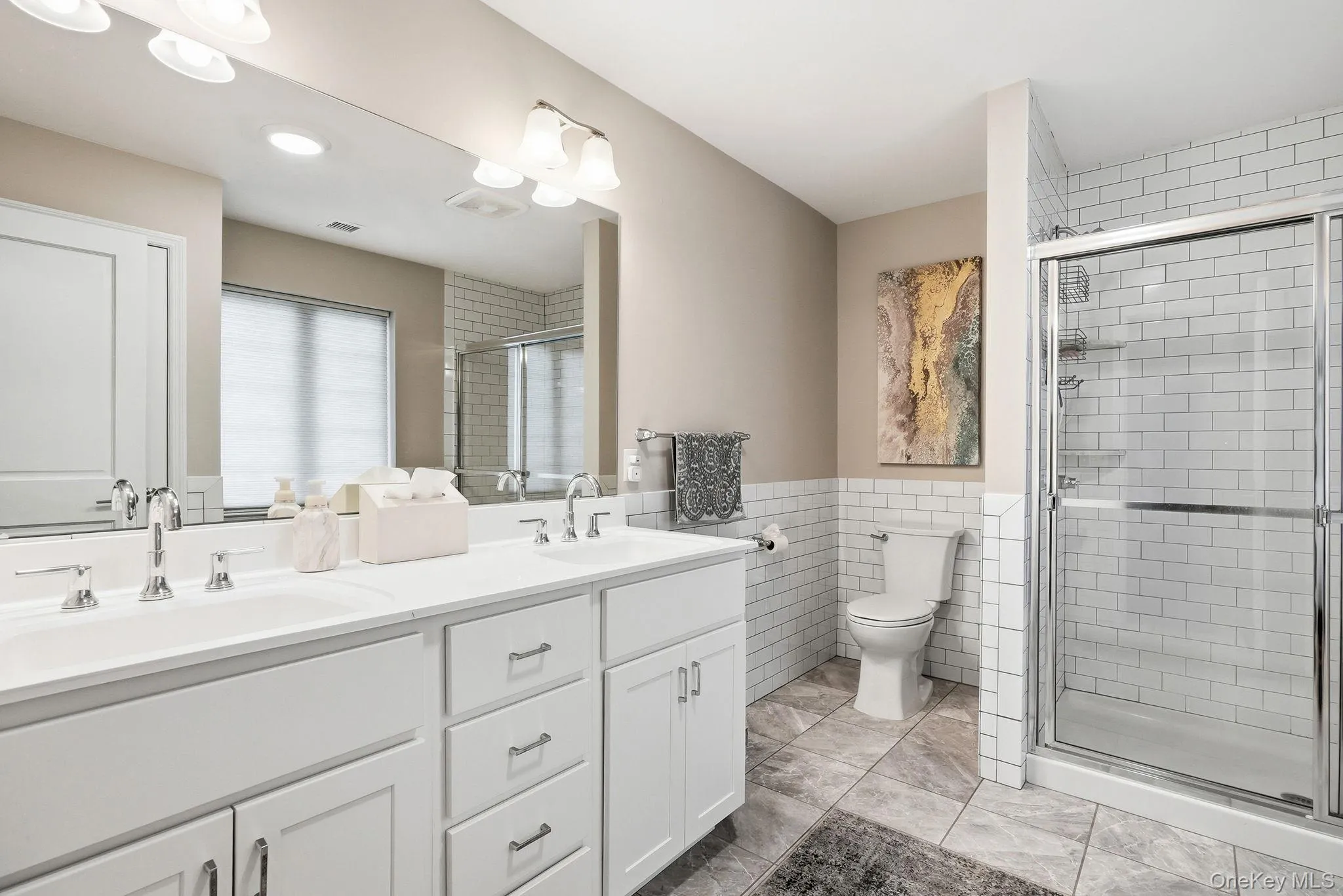 Primary bathroom featuring a double vanity with white cabinetry and chrome fixtures, subway tile wainscoting, a glass-enclosed shower with subway tile surround, and large format floor tiling Primary bathroom featuring a double vanity with white cabinetry and chrome fixtures, subway tile wainscoting, a glass-enclosed shower with subway tile surround, and large format floor tiling
