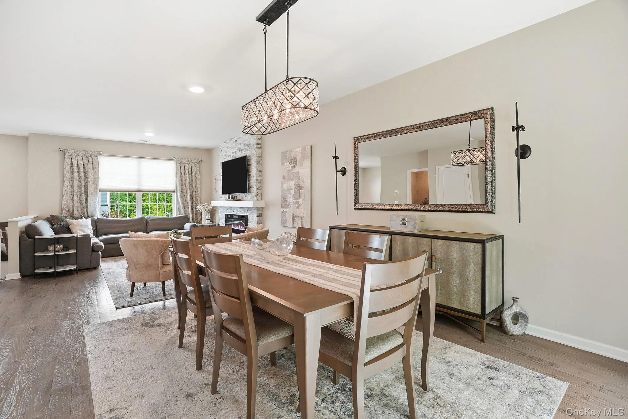 Open-concept living and dining areas featuring wood-finish flooring, a contemporary chandelier, a built-in stone-clad fireplace, and multiple windows providing natural illumination Open-concept living and dining areas featuring wood-finish flooring, a contemporary chandelier, a built-in stone-clad fireplace, and multiple windows providing natural illumination