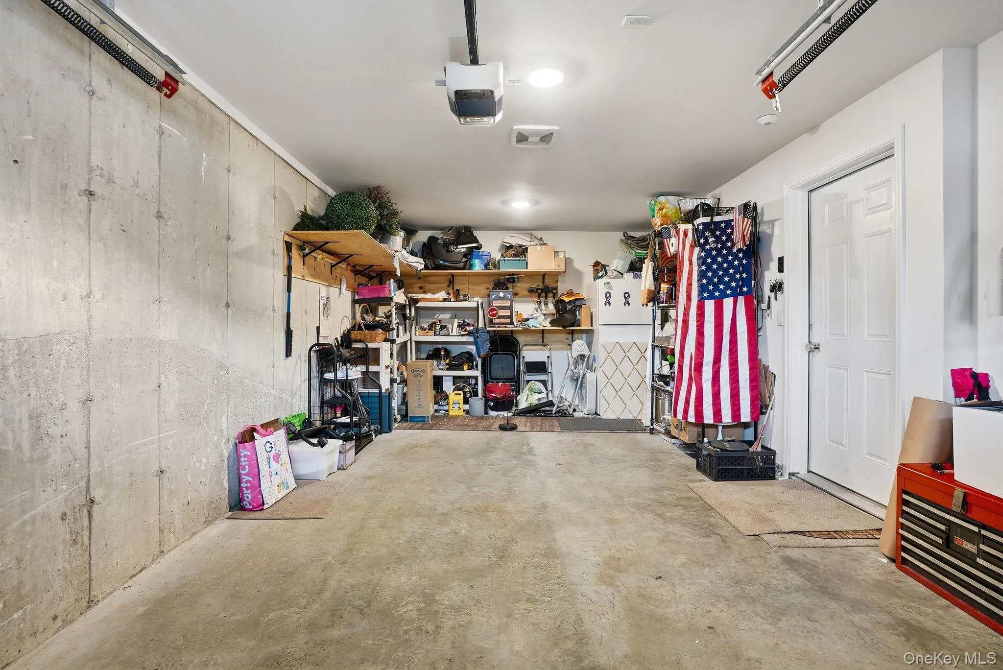 Spacious garage featuring a concrete floor, exposed concrete wall, overhead garage door opener, recessed lighting, and a white interior door Spacious garage featuring a concrete floor, exposed concrete wall, overhead garage door opener, recessed lighting, and a white interior door