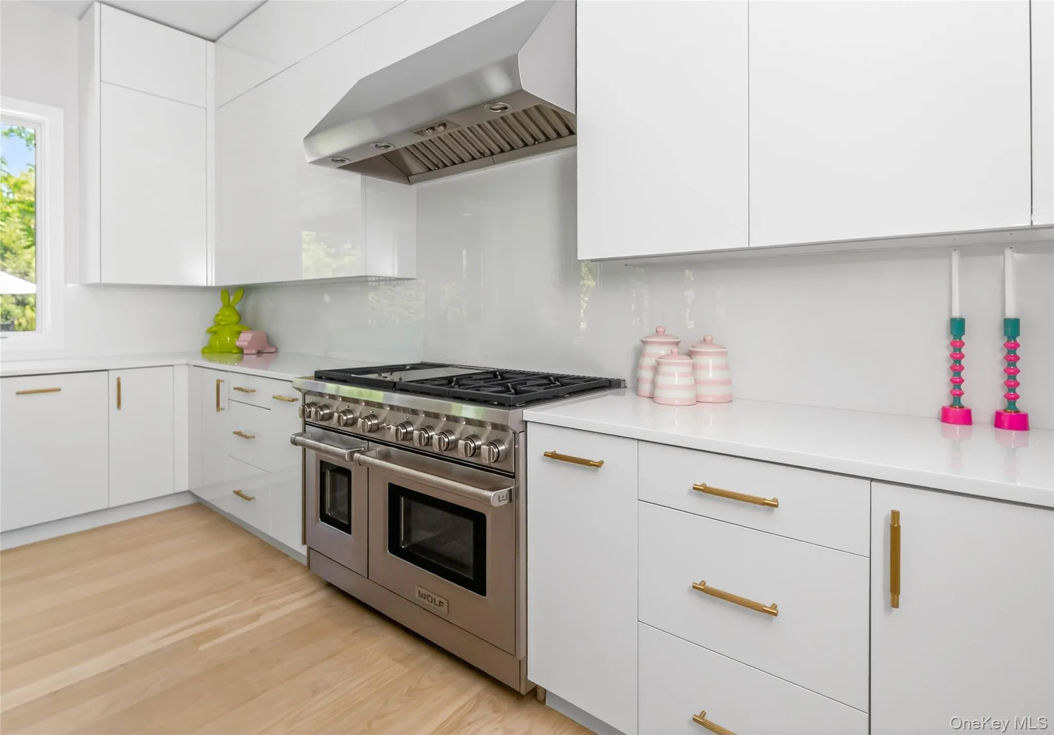 Gourmet kitchen featuring a stainless steel range with double ovens, coordinating hood vent, white cabinetry with gold-tone hardware, white countertops, and wood-finish flooring Gourmet kitchen featuring a stainless steel range with double ovens, coordinating hood vent, white cabinetry with gold-tone hardware, white countertops, and wood-finish flooring