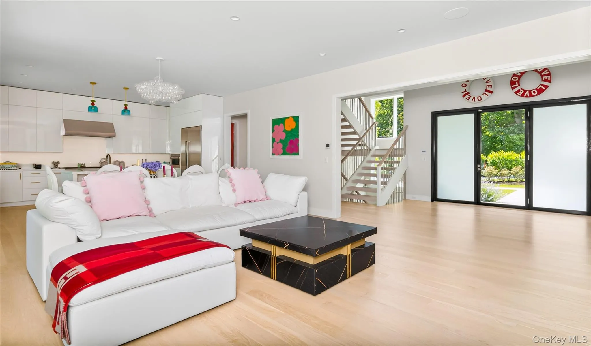 Open-concept living space with light wood-finish flooring, a contemporary kitchen featuring white cabinetry and stainless steel appliances, a modern staircase with open risers, and large glass sliding doors providing access to the exterior Open-concept living space with light wood-finish flooring, a contemporary kitchen featuring white cabinetry and stainless steel appliances, a modern staircase with open risers, and large glass sliding doors providing access to the exterior