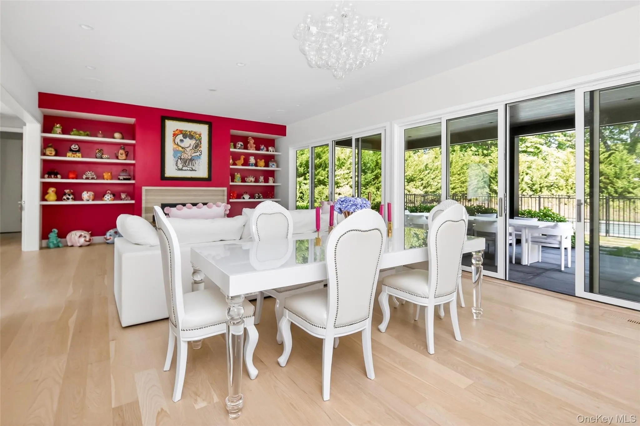 Spacious room featuring light wood-finish flooring, a contemporary chandelier, and a prominent red accent wall with built-in shelving flanking a fireplace Spacious room featuring light wood-finish flooring, a contemporary chandelier, and a prominent red accent wall with built-in shelving flanking a fireplace