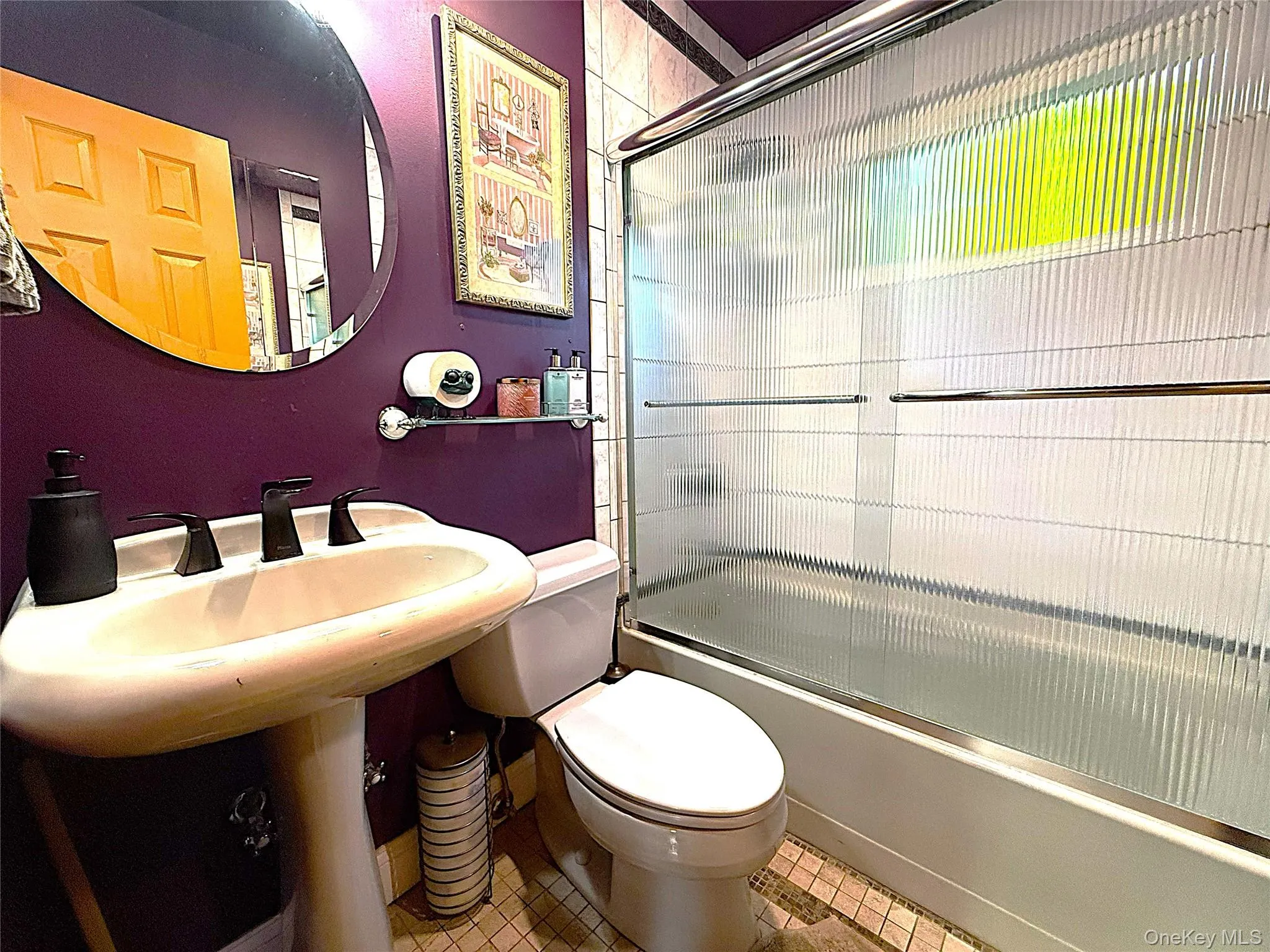 Bathroom featuring a pedestal sink with a matte black faucet, a wall-mounted oval mirror, a shower-over-tub with a fluted glass enclosure, and patterned floor tiling Bathroom featuring a pedestal sink with a matte black faucet, a wall-mounted oval mirror, a shower-over-tub with a fluted glass enclosure, and patterned floor tiling