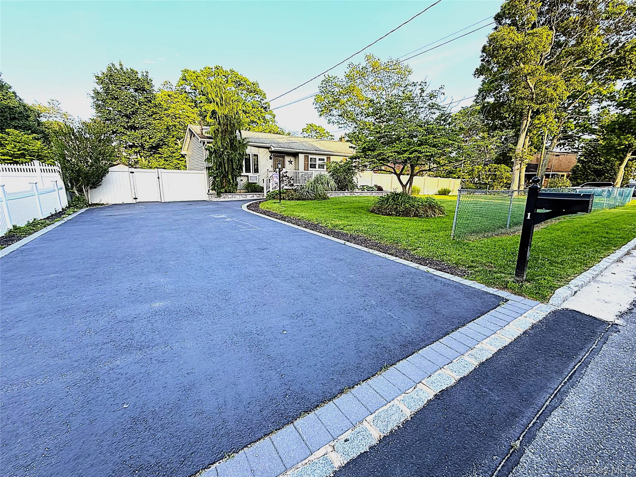 Expansive asphalt driveway featuring paver edging and a white vinyl fence with double gates Expansive asphalt driveway featuring paver edging and a white vinyl fence with double gates