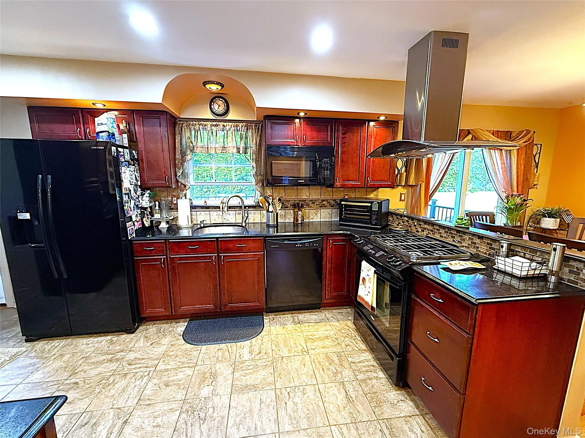 Kitchen featuring dark wood-finish cabinetry, black stone countertops, a tile backsplash, and stainless steel range hood Kitchen featuring dark wood-finish cabinetry, black stone countertops, a tile backsplash, and stainless steel range hood