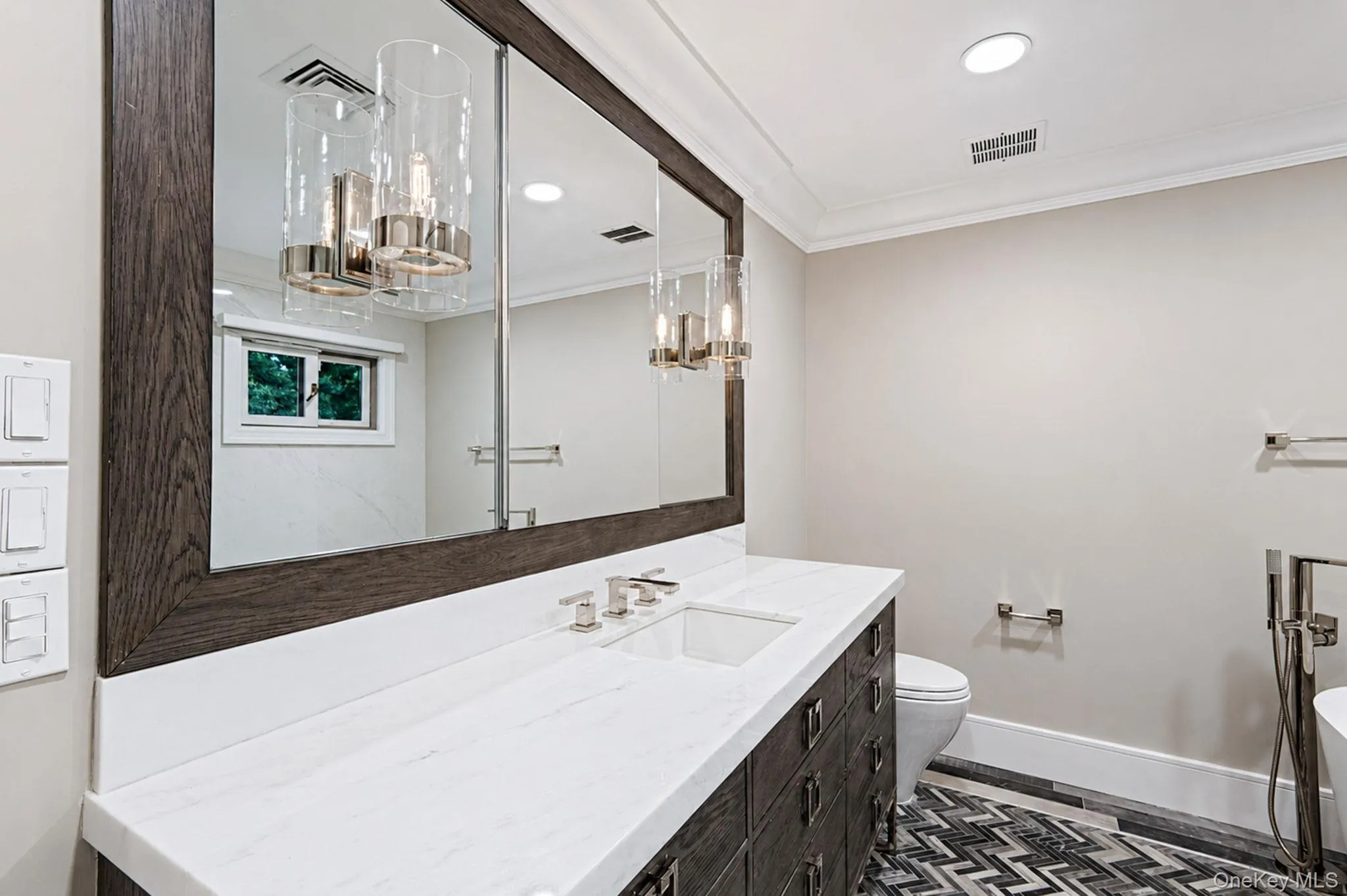 Bathroom featuring a wood-finish framed mirror with integrated sconces, a white countertop vanity with dual undermount sinks, and dark wood-finish cabinetry Bathroom featuring a wood-finish framed mirror with integrated sconces, a white countertop vanity with dual undermount sinks, and dark wood-finish cabinetry