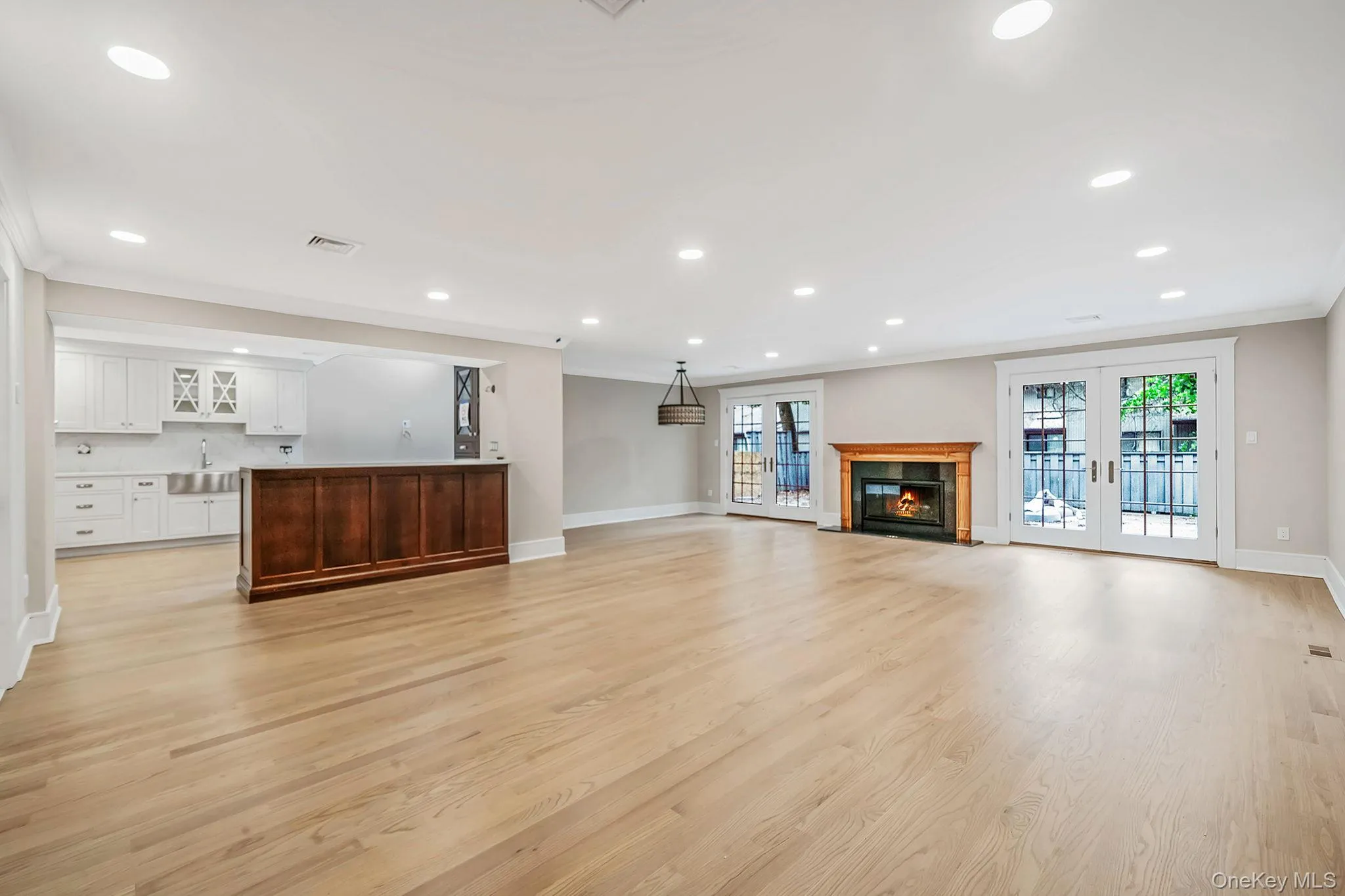 Spacious room featuring light wood-finish flooring, a built-in wet bar with dark wood cabinetry, a fireplace with a wood mantel, and two sets of double French doors Spacious room featuring light wood-finish flooring, a built-in wet bar with dark wood cabinetry, a fireplace with a wood mantel, and two sets of double French doors