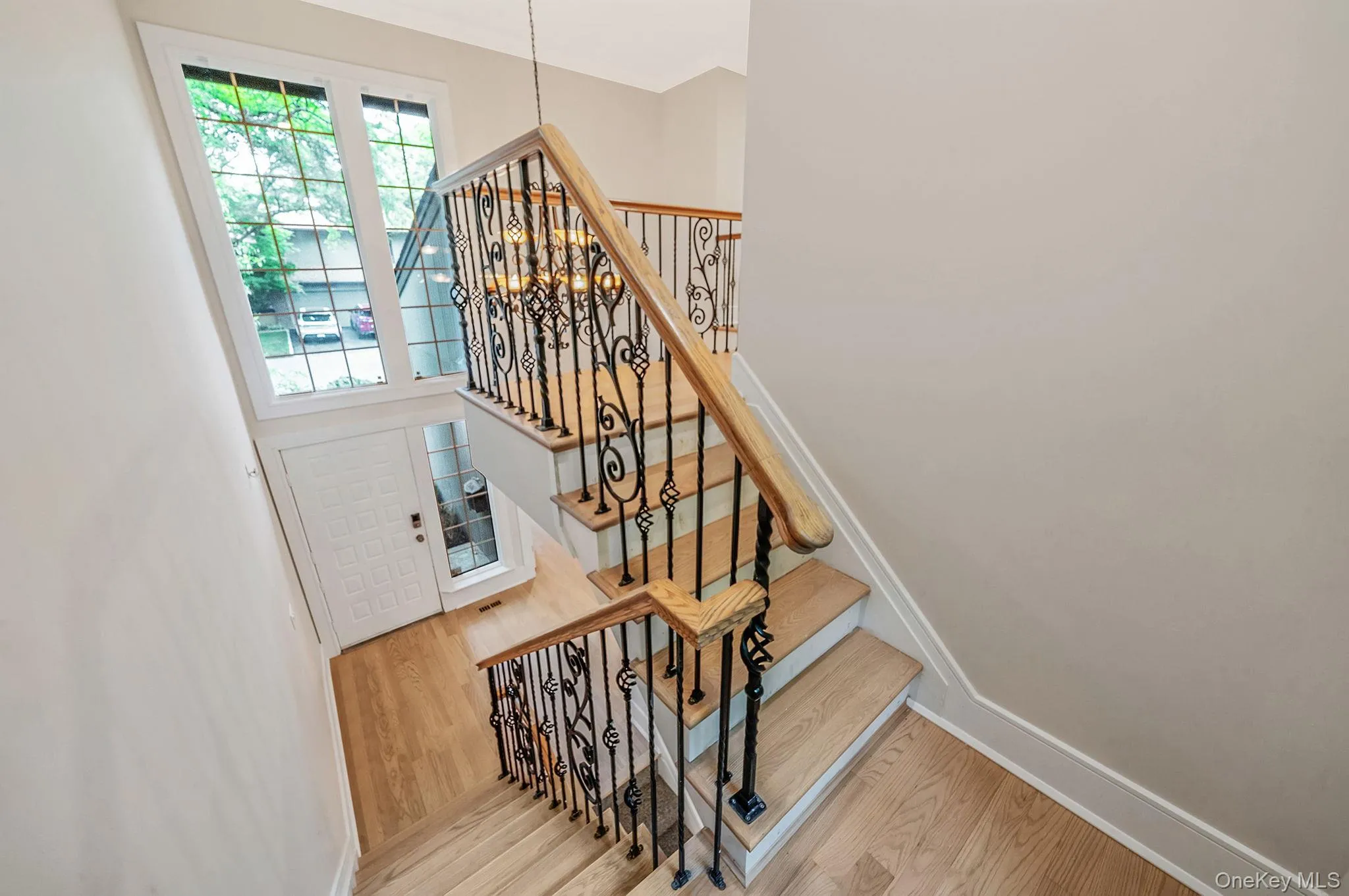 Entryway featuring a decorative white door with sidelight, light wood-finish flooring, and a grand staircase with ornate wrought iron balusters and wood handrails Entryway featuring a decorative white door with sidelight, light wood-finish flooring, and a grand staircase with ornate wrought iron balusters and wood handrails