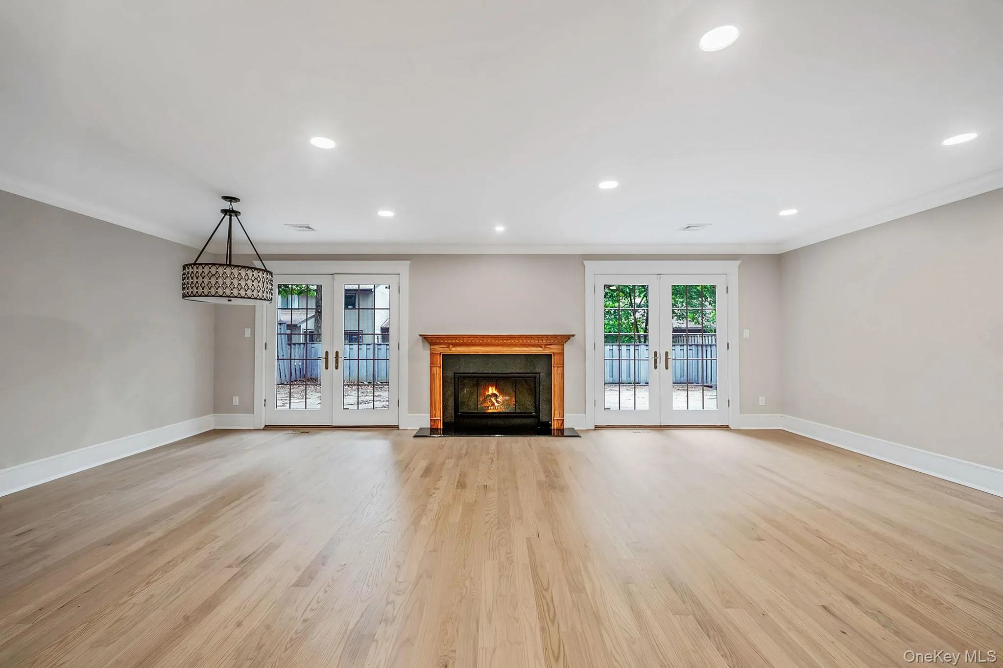 Spacious room featuring a wood-finish floor, a central fireplace with a wood mantel, two sets of French doors, recessed lighting, and a decorative chandelier Spacious room featuring a wood-finish floor, a central fireplace with a wood mantel, two sets of French doors, recessed lighting, and a decorative chandelier