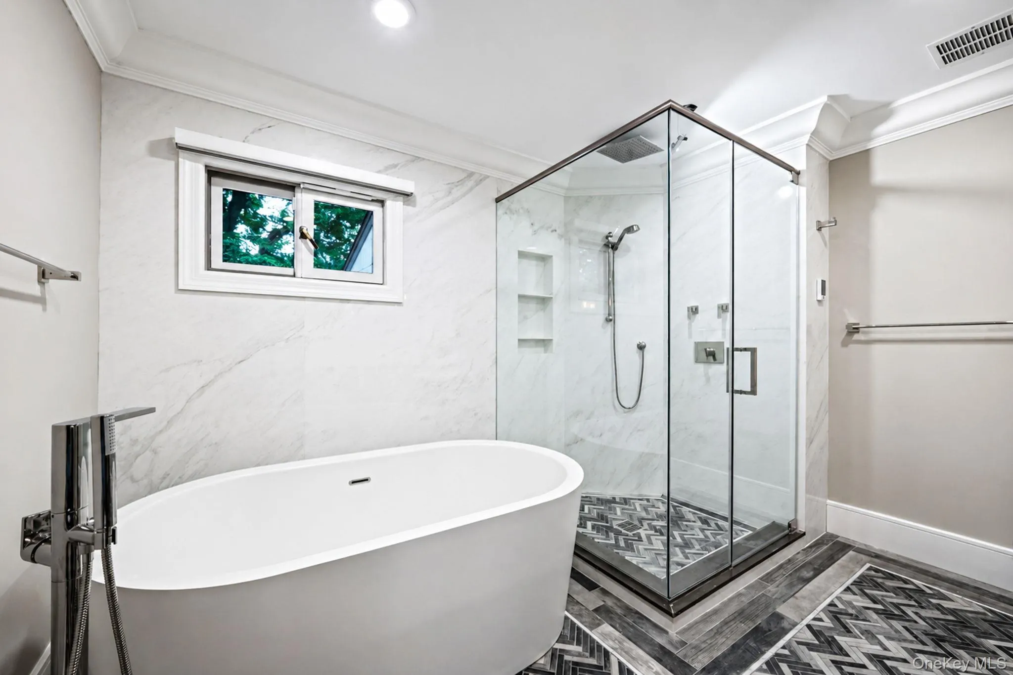 Freestanding soaking tub with chrome floor-mounted faucet, glass-enclosed shower with tiled walls and built-in niches, and chevron-patterned floor tiling Freestanding soaking tub with chrome floor-mounted faucet, glass-enclosed shower with tiled walls and built-in niches, and chevron-patterned floor tiling