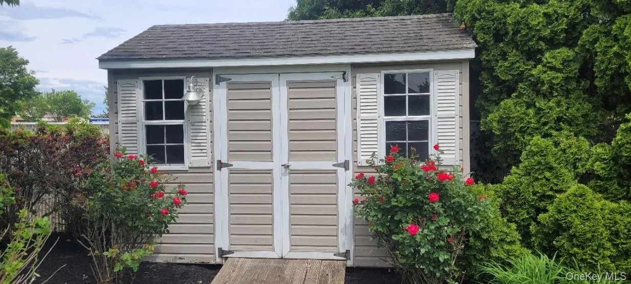 Exterior storage shed with vinyl siding, shingled roof, double entry doors, window with louvered shutters, and exterior wall-mounted lighting Exterior storage shed with vinyl siding, shingled roof, double entry doors, window with louvered shutters, and exterior wall-mounted lighting