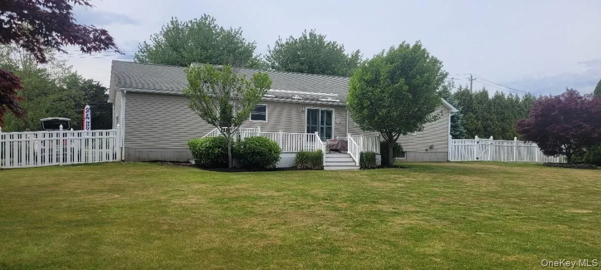 Expansive rear lawn featuring a white picket fence, mature trees, and a ground-level deck with white railings and sliding glass door access Expansive rear lawn featuring a white picket fence, mature trees, and a ground-level deck with white railings and sliding glass door access