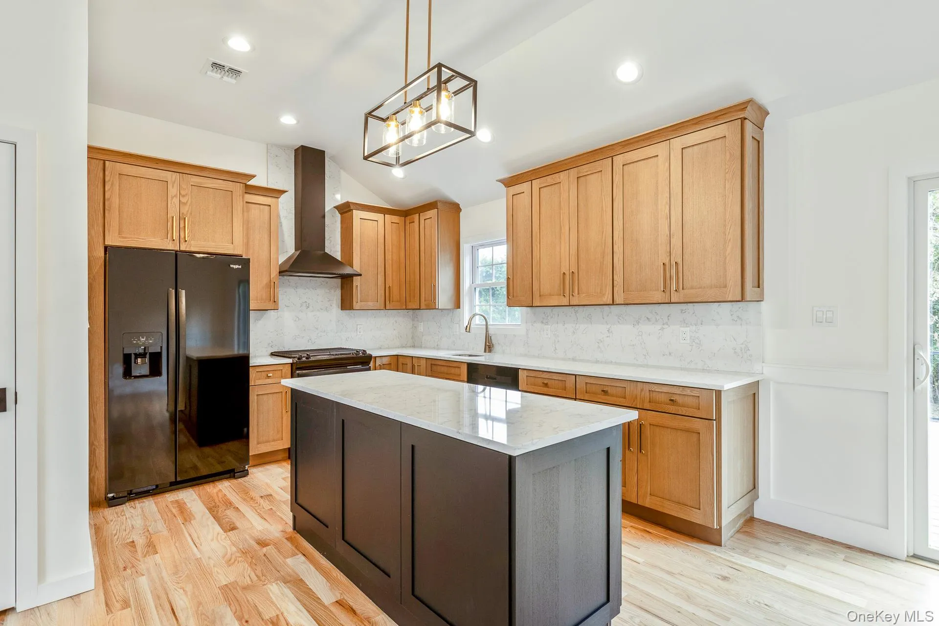 Kitchen featuring black refrigerator with ice dispenser, light stone counters, tasteful backsplash, a center island, and wall chimney exhaust hood Kitchen featuring black refrigerator with ice dispenser, light stone counters, tasteful backsplash, a center island, and wall chimney exhaust hood