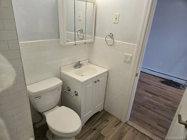 Bathroom featuring white subway tile wainscoting, a white vanity with integrated sink, a mirrored medicine cabinet, and wood-finish flooring Bathroom featuring white subway tile wainscoting, a white vanity with integrated sink, a mirrored medicine cabinet, and wood-finish flooring