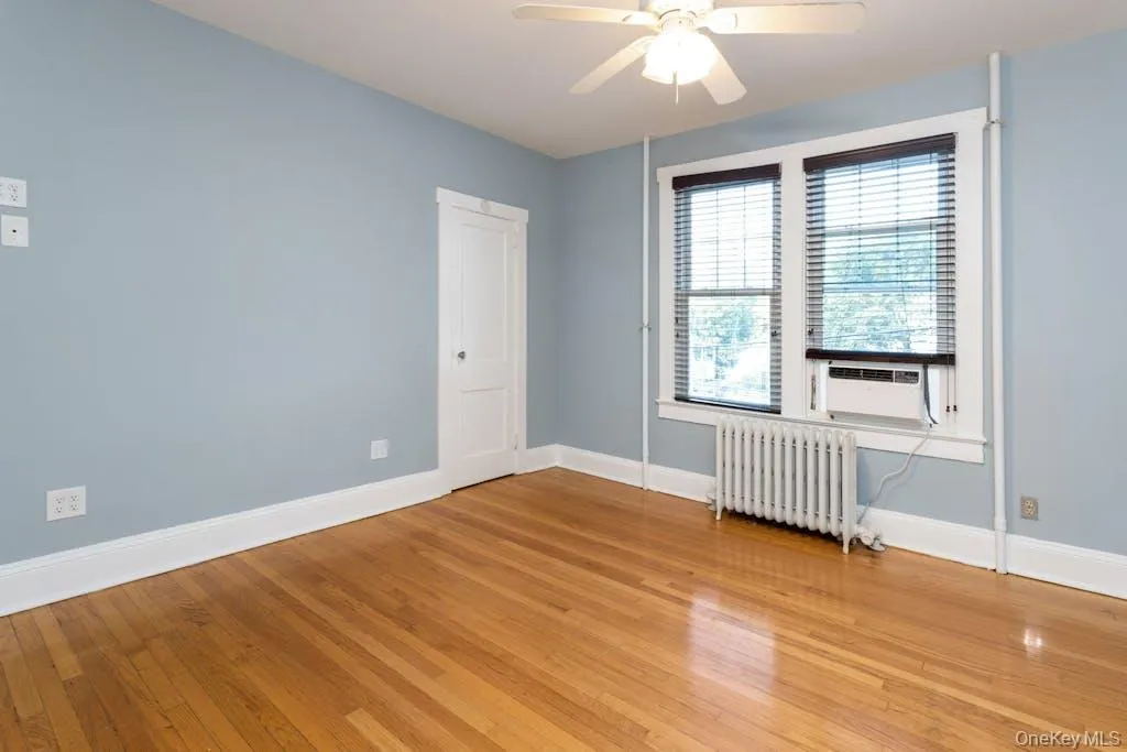 Empty room featuring ceiling fan, radiator heating unit, light hardwood / wood-style flooring, and cooling unit Empty room featuring ceiling fan, radiator heating unit, light hardwood / wood-style flooring, and cooling unit