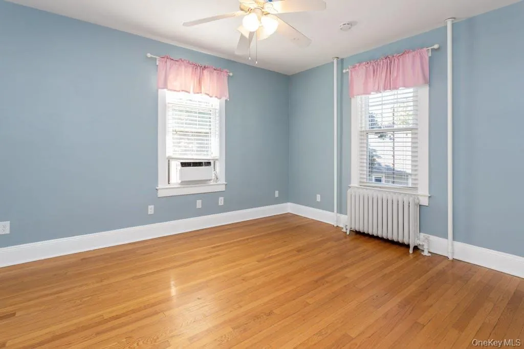 Unfurnished room with hardwood / wood-style floors, ceiling fan, a wealth of natural light, and radiator heating unit Unfurnished room with hardwood / wood-style floors, ceiling fan, a wealth of natural light, and radiator heating unit