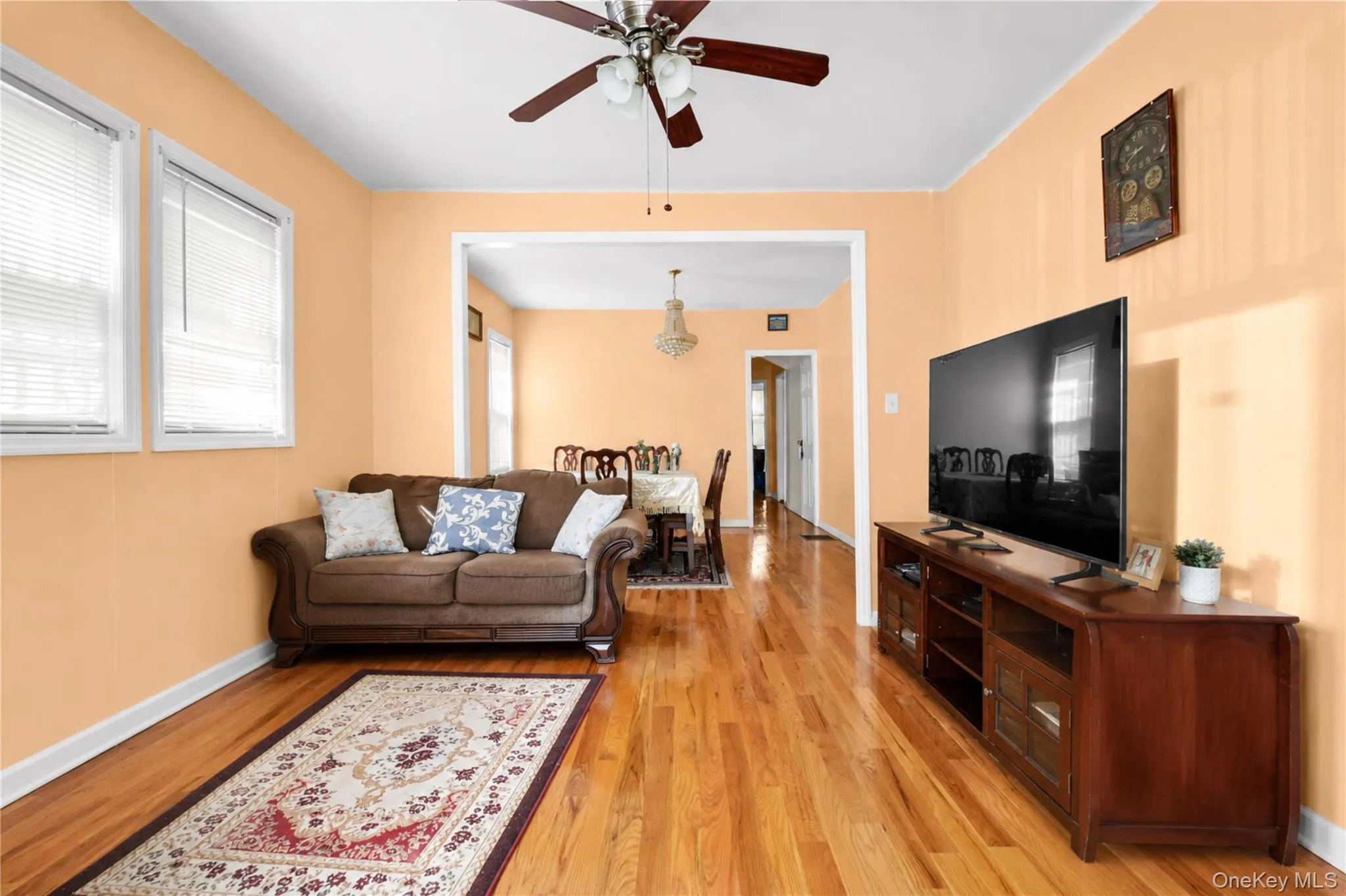 Living area featuring wood-finish flooring, two windows with blinds, a ceiling fan, and an expansive archway leading to a dining area Living area featuring wood-finish flooring, two windows with blinds, a ceiling fan, and an expansive archway leading to a dining area