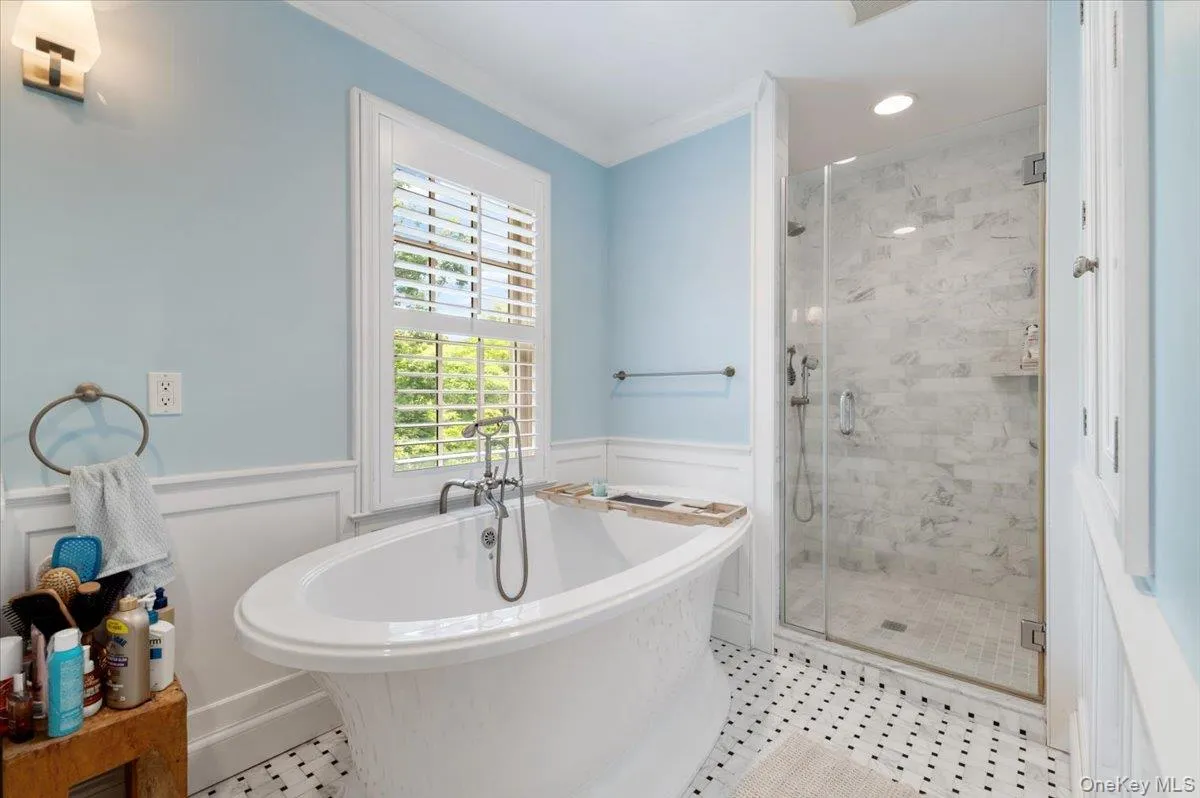 Freestanding soaking tub with chrome fixtures, glass-enclosed shower with marble-look tile surround, white wainscoting, louvered window treatments, and patterned floor tile Freestanding soaking tub with chrome fixtures, glass-enclosed shower with marble-look tile surround, white wainscoting, louvered window treatments, and patterned floor tile