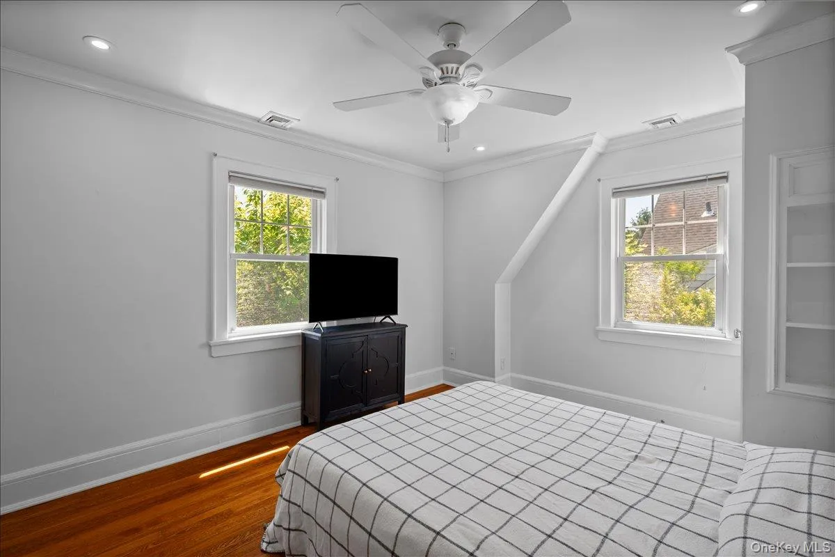 Primary bedroom featuring hardwood flooring, two windows with white trim, a ceiling fan, recessed lighting, and built-in shelving Primary bedroom featuring hardwood flooring, two windows with white trim, a ceiling fan, recessed lighting, and built-in shelving