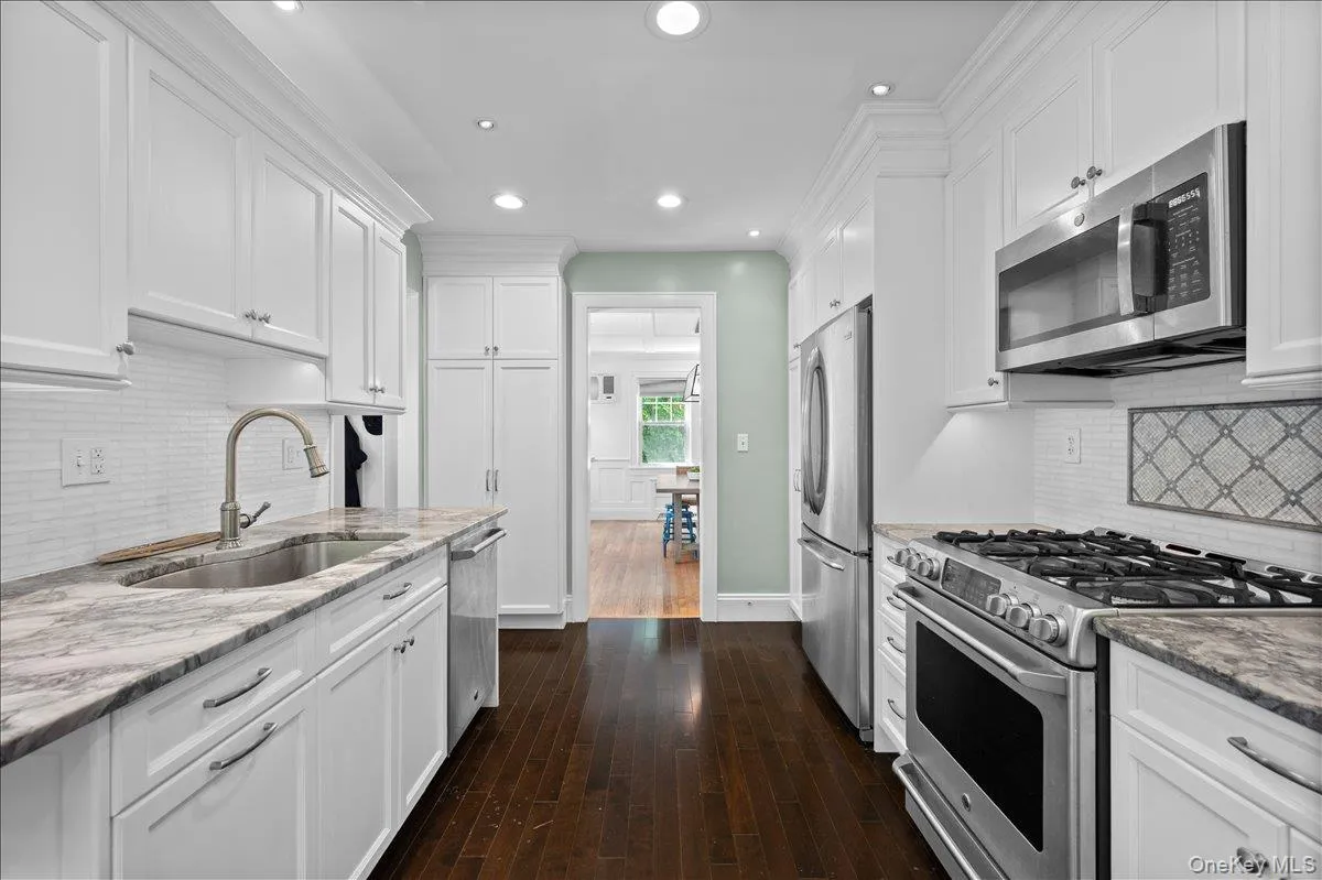 Galley kitchen featuring white shaker-style cabinetry, stainless steel appliances, dark wood-finish flooring, and light-colored countertops Galley kitchen featuring white shaker-style cabinetry, stainless steel appliances, dark wood-finish flooring, and light-colored countertops