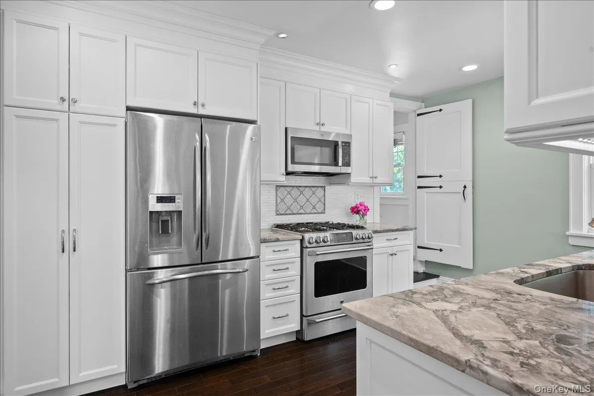 Kitchen featuring extensive white cabinetry, stainless steel appliances, dark wood-finish flooring, and light-toned stone countertops Kitchen featuring extensive white cabinetry, stainless steel appliances, dark wood-finish flooring, and light-toned stone countertops