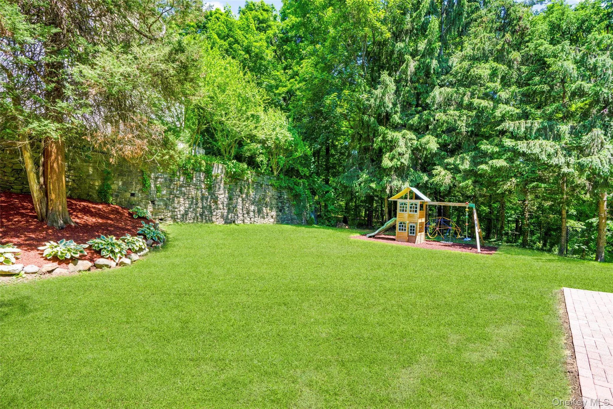 Expansive backyard featuring a lush green lawn, mature trees, a stone retaining wall, and a paved brick patio area Expansive backyard featuring a lush green lawn, mature trees, a stone retaining wall, and a paved brick patio area