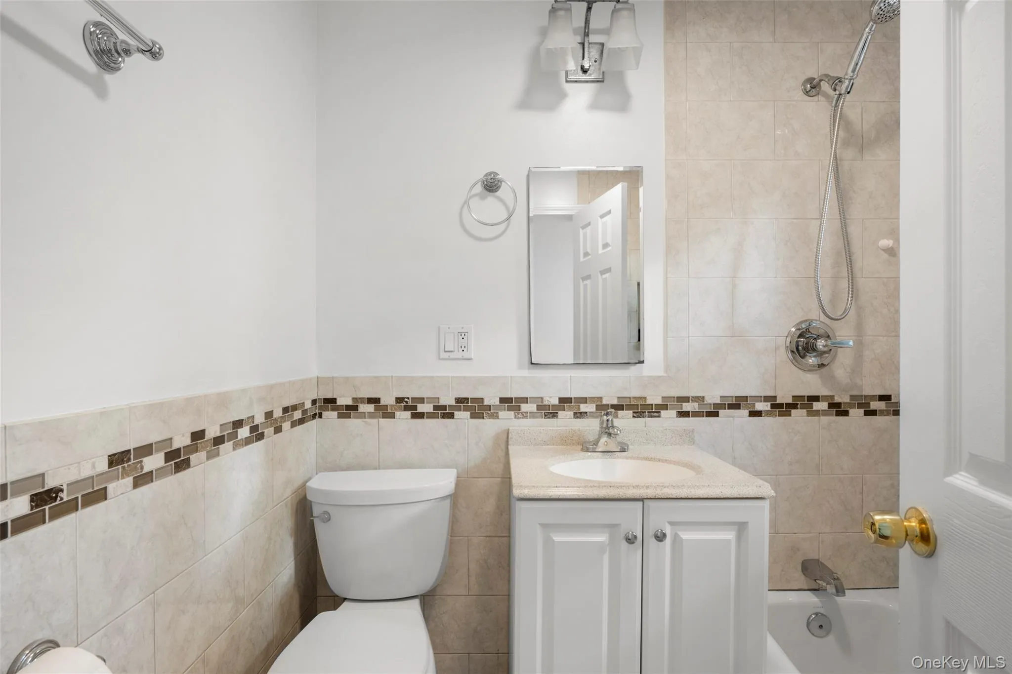 Bathroom featuring a white vanity with a light-colored countertop, ceramic tile walls with a decorative mosaic accent, and polished chrome fixtures including a showerhead and faucet Bathroom featuring a white vanity with a light-colored countertop, ceramic tile walls with a decorative mosaic accent, and polished chrome fixtures including a showerhead and faucet