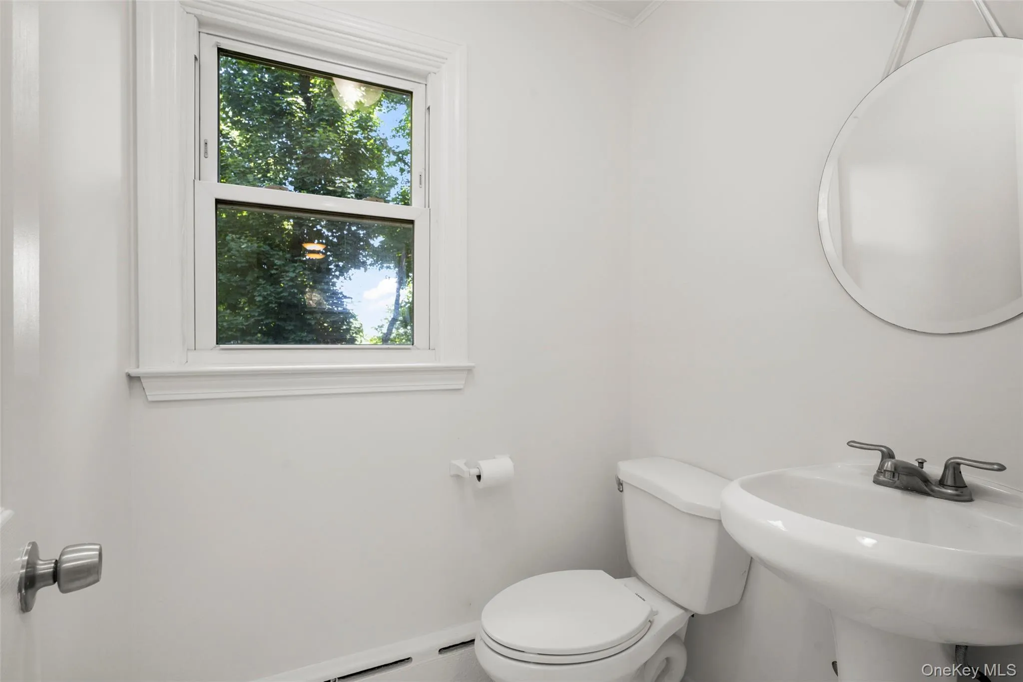 Powder room featuring a single-pane window with detailed trim, pedestal sink with chrome faucet, and a round mirror Powder room featuring a single-pane window with detailed trim, pedestal sink with chrome faucet, and a round mirror