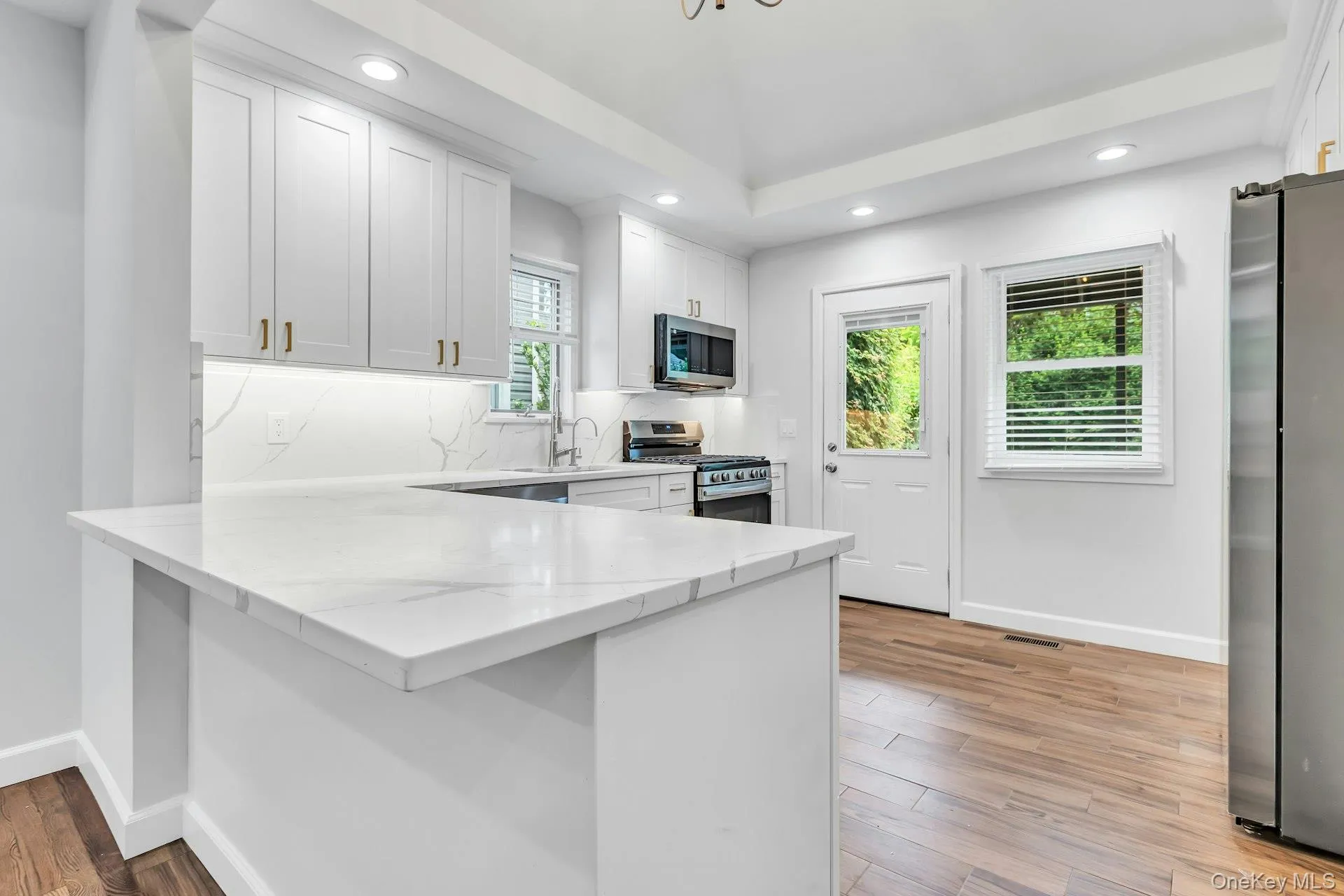 Modern kitchen featuring white shaker cabinetry, quartz countertops with a matching backsplash, stainless steel appliances, and wood-finish flooring Modern kitchen featuring white shaker cabinetry, quartz countertops with a matching backsplash, stainless steel appliances, and wood-finish flooring
