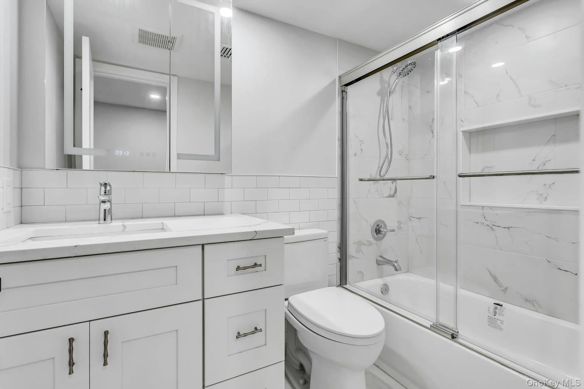 Bathroom featuring a white subway tile backsplash, white vanity with integrated sink, chrome faucet, and an illuminated mirrored medicine cabinet Bathroom featuring a white subway tile backsplash, white vanity with integrated sink, chrome faucet, and an illuminated mirrored medicine cabinet