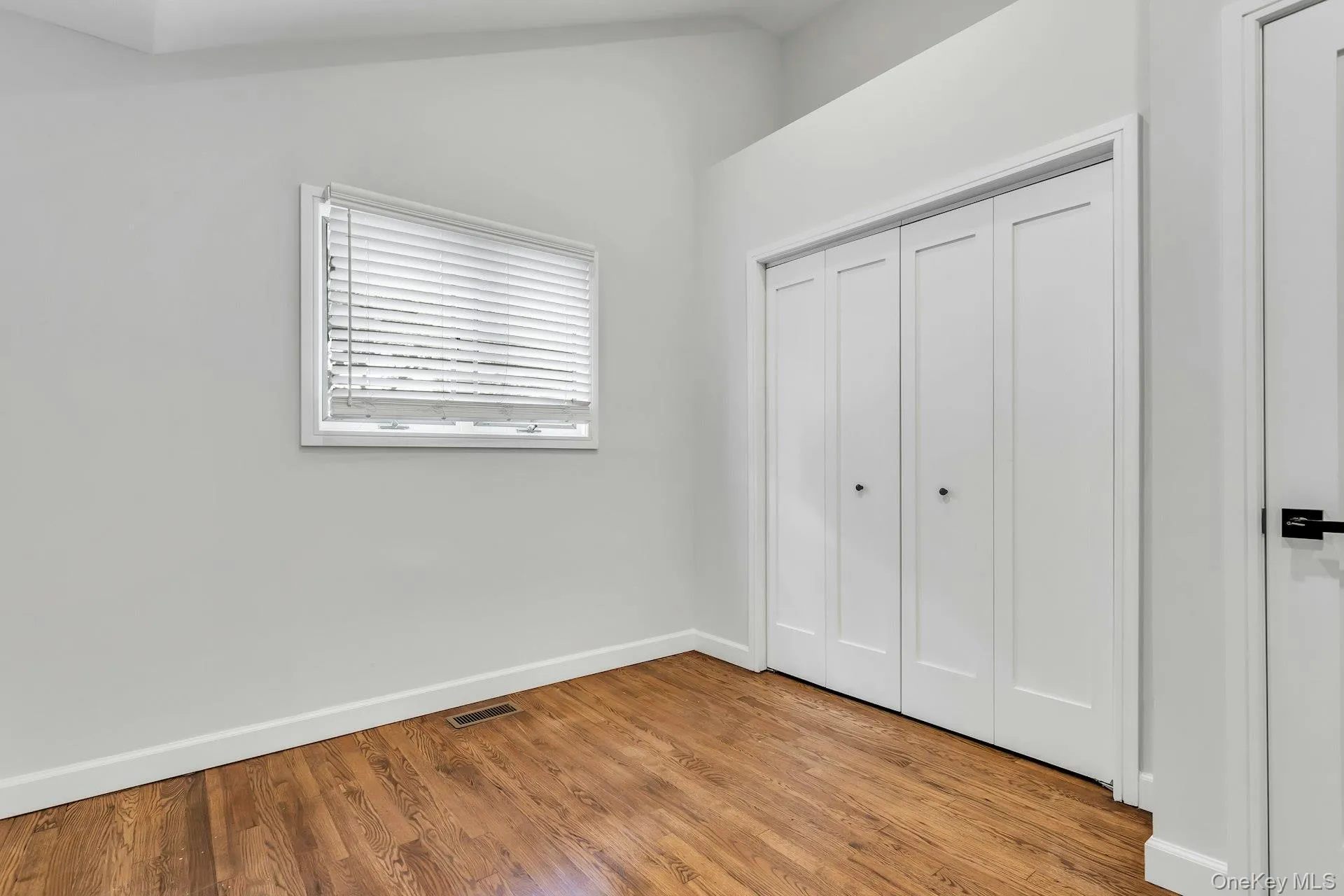 Wood-finish flooring complemented by white baseboards, a single window with white blinds, and bi-fold closet doors with black hardware Wood-finish flooring complemented by white baseboards, a single window with white blinds, and bi-fold closet doors with black hardware