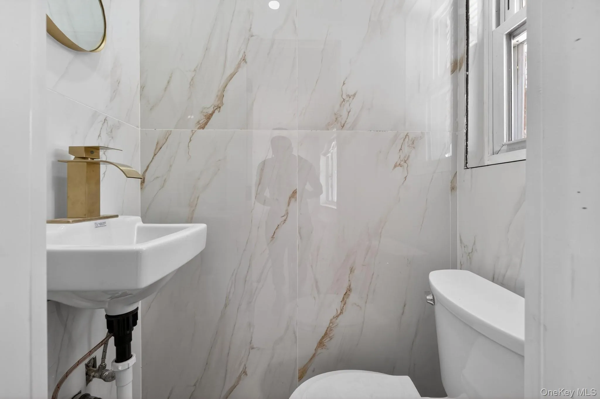 Bathroom featuring full-height marbled wall tiling, a contemporary white ceramic wall-mounted sink, and a modern gold-finish waterfall faucet Bathroom featuring full-height marbled wall tiling, a contemporary white ceramic wall-mounted sink, and a modern gold-finish waterfall faucet