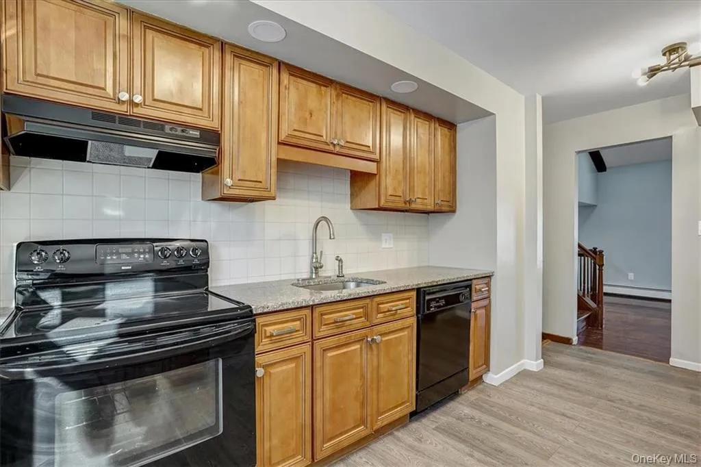 Kitchen featuring black appliances, light stone countertops, under cabinet range hood, brown cabinetry, and light wood-style flooring Kitchen featuring black appliances, light stone countertops, under cabinet range hood, brown cabinetry, and light wood-style flooring
