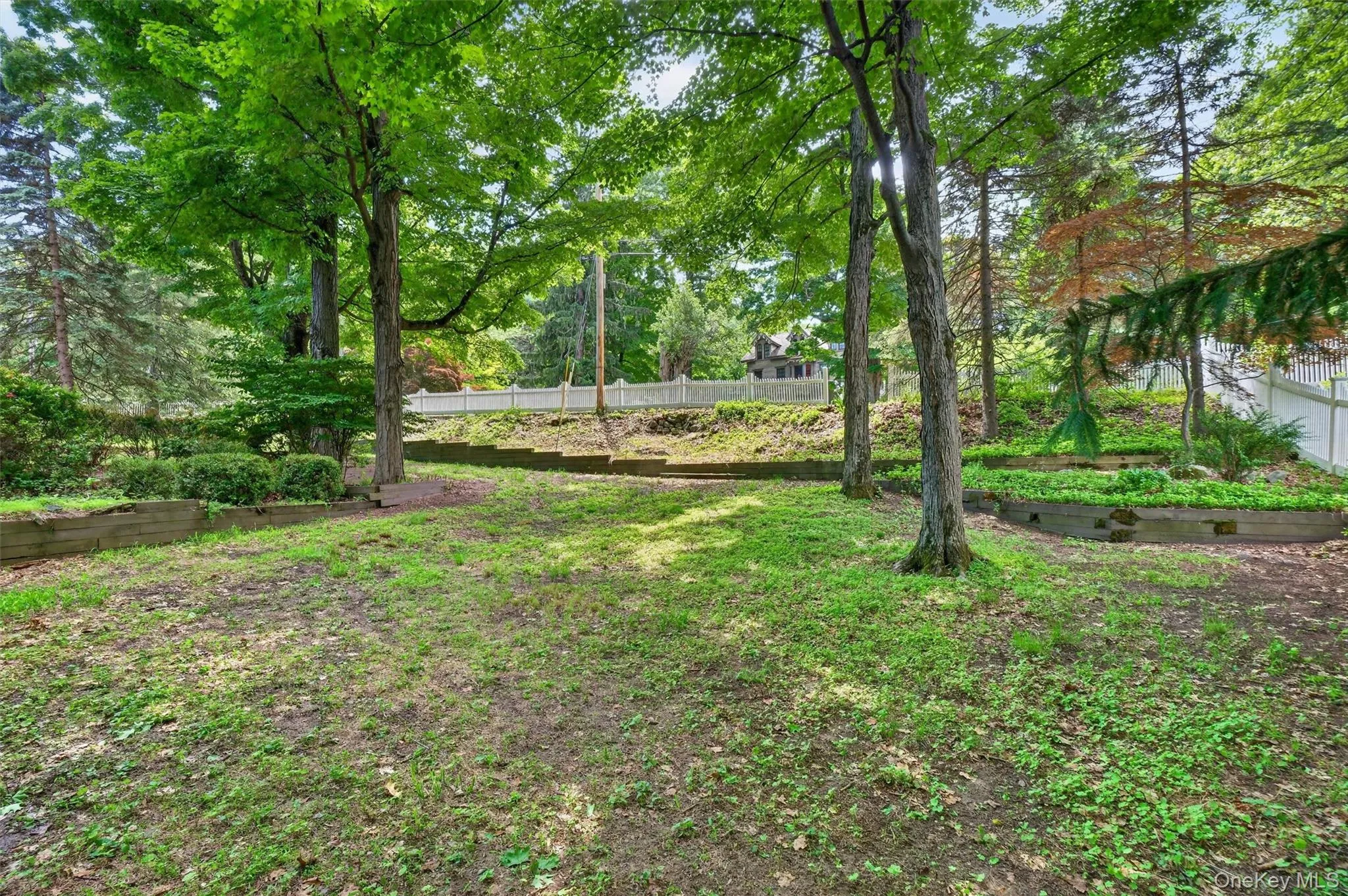 Tiered landscaping featuring mature trees, established plantings, and a white picket fence Tiered landscaping featuring mature trees, established plantings, and a white picket fence