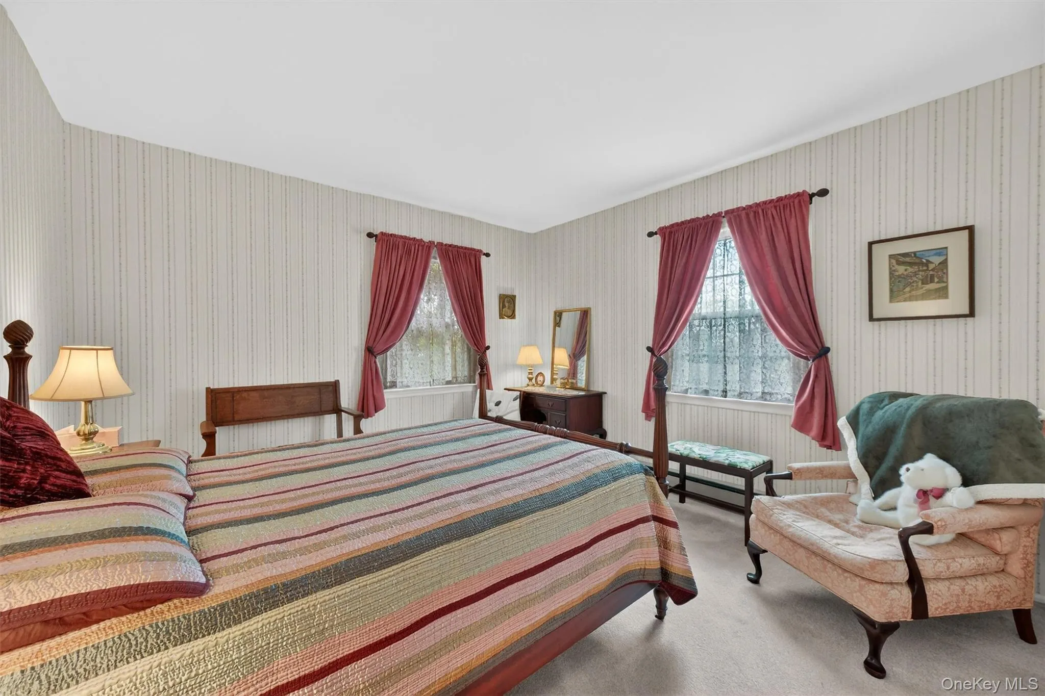 Spacious bedroom featuring light-colored striped wallpaper, two windows with red draperies, and light-toned carpeting Spacious bedroom featuring light-colored striped wallpaper, two windows with red draperies, and light-toned carpeting
