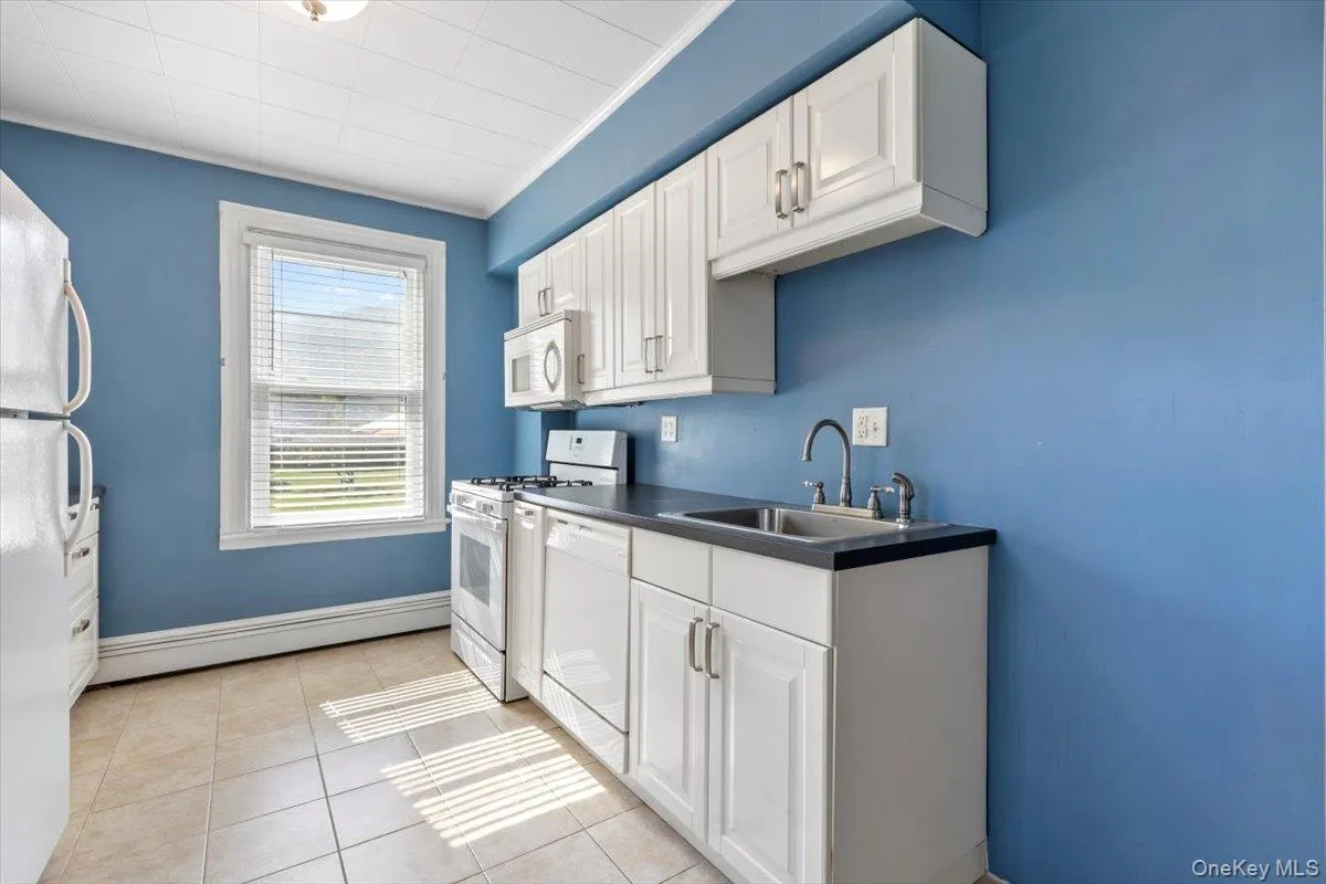 Kitchen featuring a sink, white cabinetry, white appliances, dark countertops, and baseboard heating Kitchen featuring a sink, white cabinetry, white appliances, dark countertops, and baseboard heating