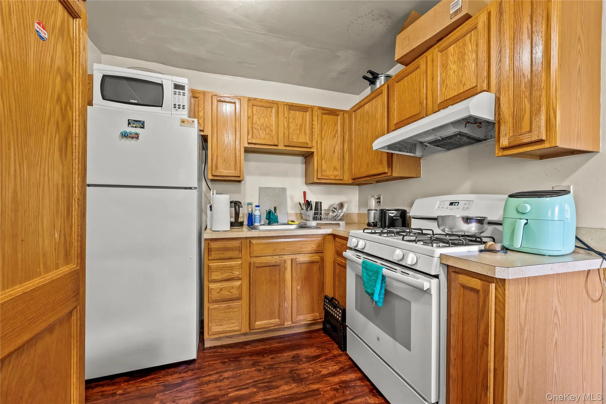 Kitchen featuring white appliances, light countertops, under cabinet range hood, dark wood finished floors, and brown cabinets Kitchen featuring white appliances, light countertops, under cabinet range hood, dark wood finished floors, and brown cabinets
